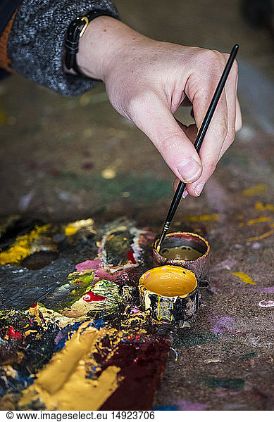 High angle close up of human hand dipping paintbrush into small pot of yellow oil paint.
