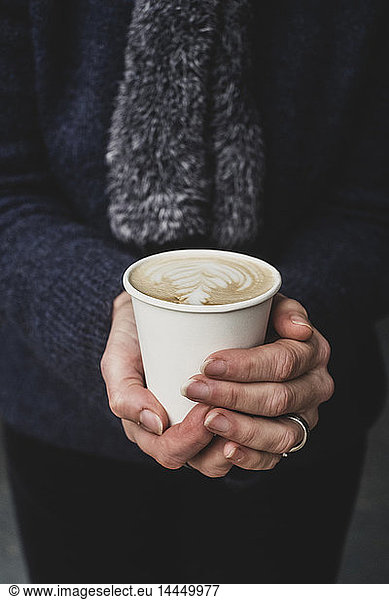 High angle close up of person holding paper cup with cafe latte topped with intricate foam pattern.