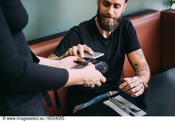 High angle close up of tattooed bearded man sitting in a bar  using mobile phone to pay.