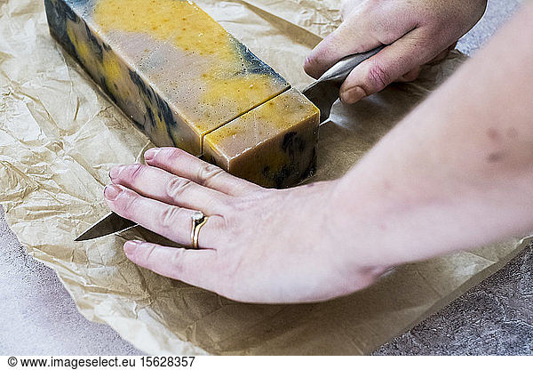 High angle close up person cutting yellow and black homemade bar of soap with kitchen knife.