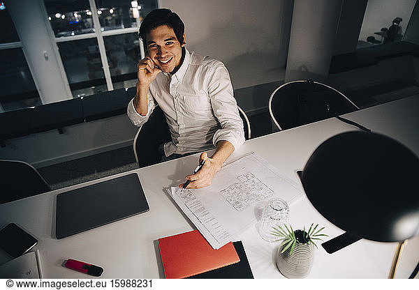 High angle portrait of smiling businessman sitting with blueprint at desk while working late in office