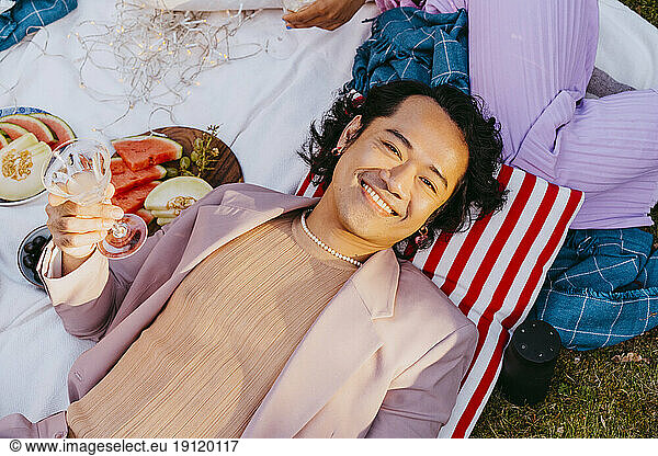 High angle portrait of smiling gay man holding wineglass while lying down in back yard