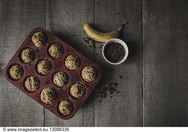 High angle view of baked muffins in baking sheet by chocolate chips and banana on wooden table at home