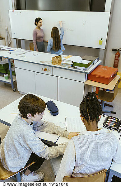 High angle view of boy helping male friend while sitting on desk in classroom