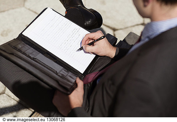 High angle view of businessman writing on document while sitting on bench