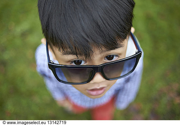 High angle view of cute boy wearing oversized black sunglasses while standing on field at park