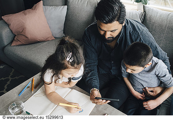 High angle view of father showing mobile phone to son while daughter drawing in book at home