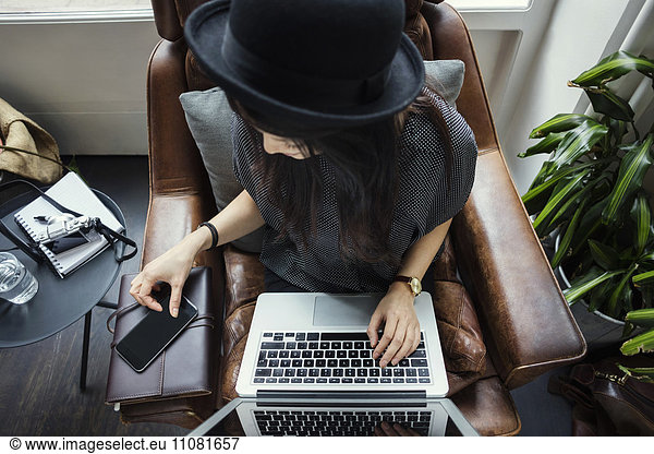 High angle view of female blogger using phone and laptop while sitting on chair