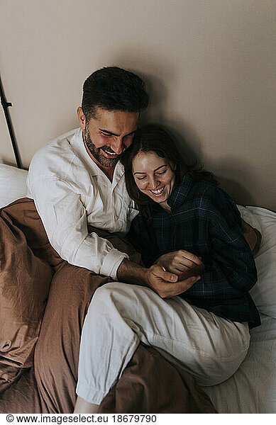 High angle view of happy couple sharing smart phone while sitting on bed at home