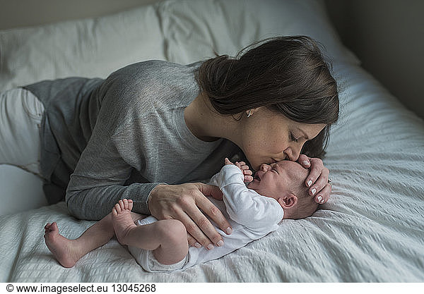 High angle view of mother kissing crying newborn baby boy on forehead in bed