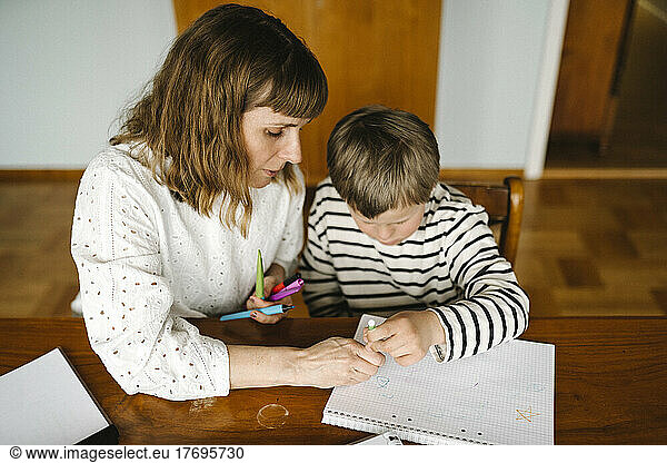 High angle view of mother teaching son with down syndrome to draw in book at table