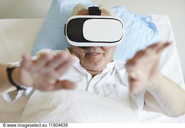 High angle view of senior man gesturing while using VR glasses on bed in hospital ward