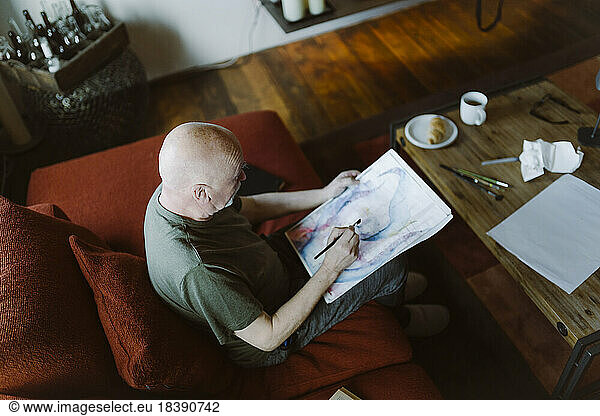 High angle view of senior man painting face over paper while sitting on sofa at home