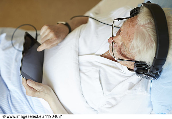 High angle view of senior man wearing headphones while touching digital tablet in hospital ward