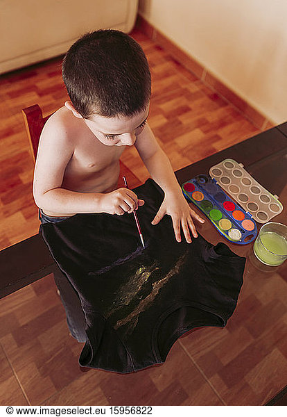 High angle view of shirtless boy drawing on t-shirt with watercolor paints at home