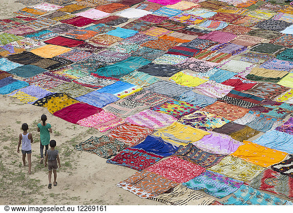 High angle view of three children standing on a field with a large number of colourful cotton fabrics spread out for drying.