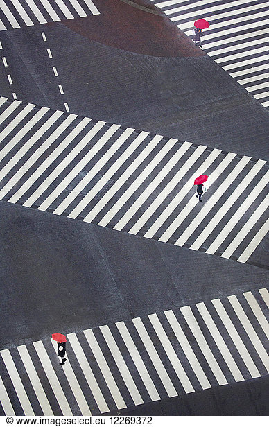 High angle view of three people carrying bright red umbrellas walking across urban street at pedestrian crossings.