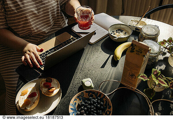 High angle view of woman holding drinking glass while using laptop with breakfast on table at home