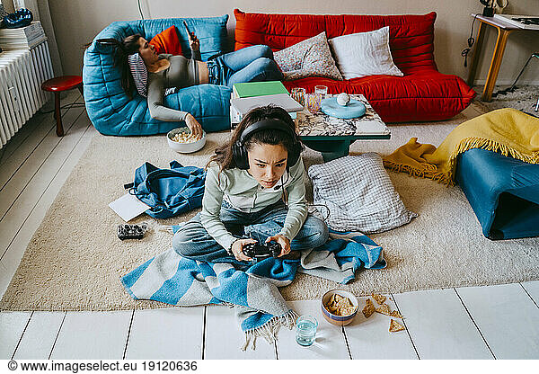 High angle view of woman playing video game while friend using smart phone on sofa at home