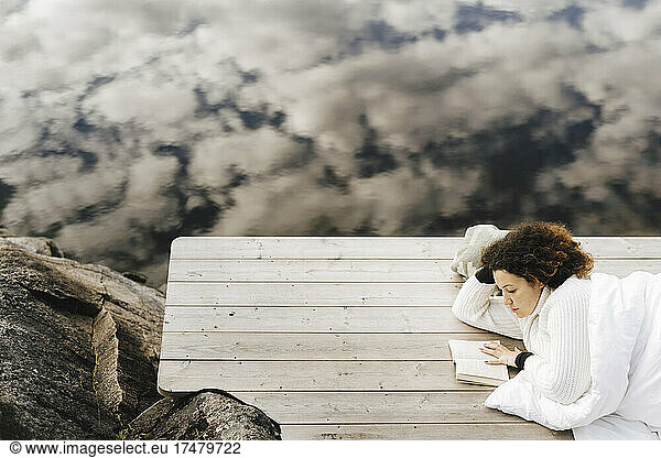 High angle view of woman reading book while lying cloud's reflection on lake