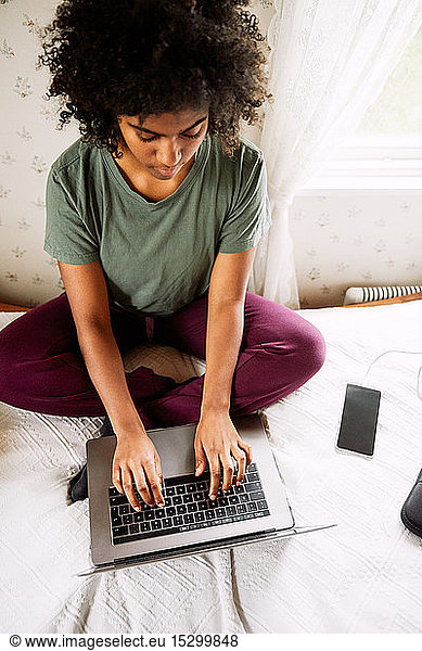 High angle view of young woman with curly hair using laptop while sitting on bed at home