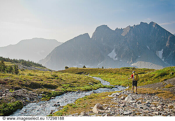 Hiker wearing backpack hikes beside tranquil stream in alpine meadow.