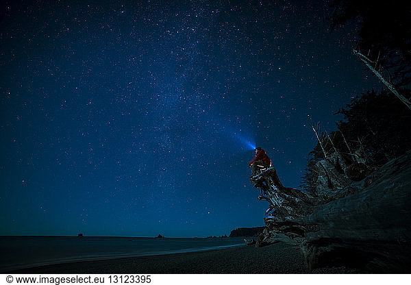 Hiker wearing headlamp sitting on tree trunk at La Push beach against sky at night