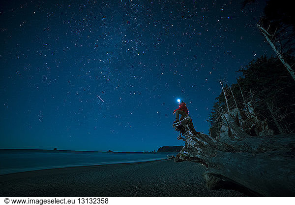 Hiker wearing headlamp sitting on tree trunk at La Push beach against star field