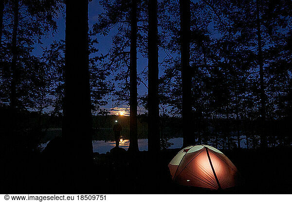 Hiker with a flashlight is staying near lake shore at his tent.