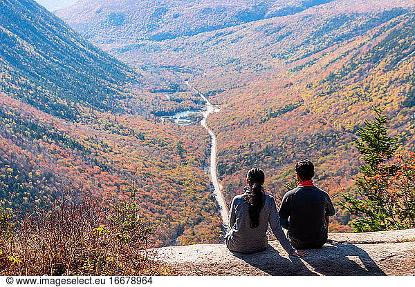 Hiking couple sitting on mountain ledge looking out over valley in NH.