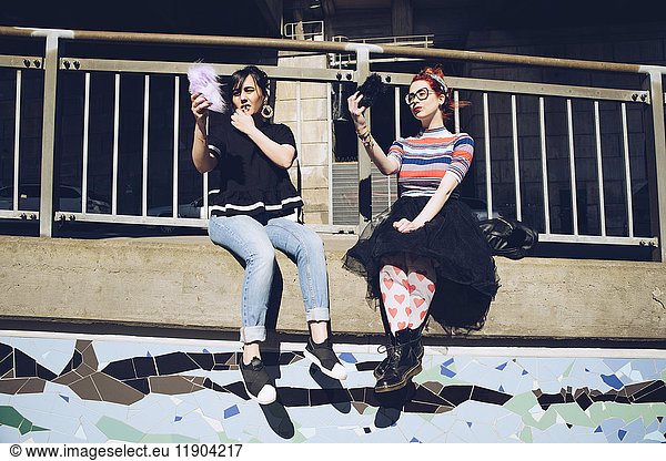 Hipster female friends taking selfie with mobile phones while sitting on wall against railing