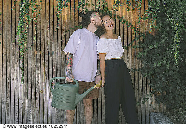 Hipster man holding watering can kissing happy girlfriend in front of wooden wall