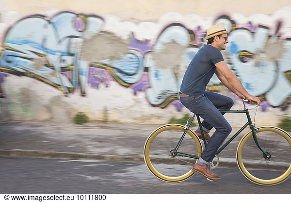 Hipster man riding bicycle on road along urban graffiti wall