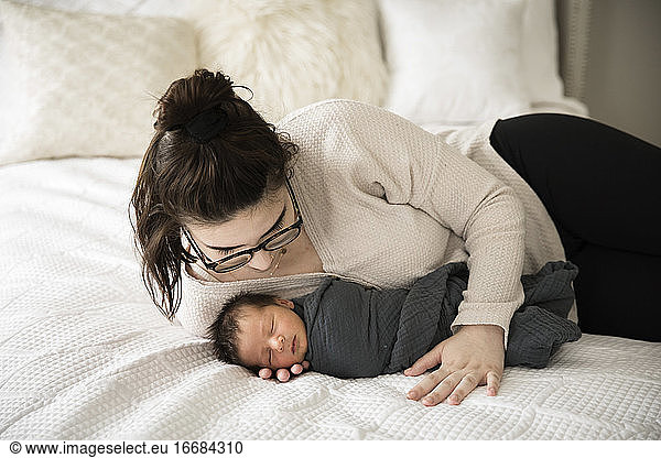 Hipster Millennial Mom Snuggles Swaddled Newborn on White Bed
