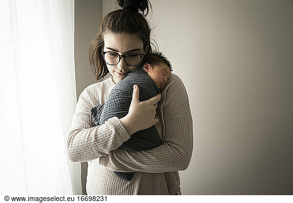 Hipster Millennial Mom Snuggles Swaddled Newborn Sohn neben dem Fenster