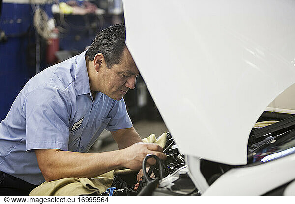Hispanic mechanic leans on a car working on the engine compartment in an auto repair shop