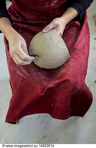 Hochformatige Nahaufnahme einer Keramikkünstlerin mit roter Schürze  die in ihrer Werkstatt sitzt und an einer Tonvase arbeitet.