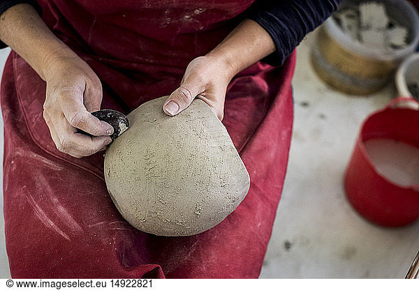 Hochformatige Nahaufnahme einer Keramikkünstlerin mit roter Schürze  die in ihrer Werkstatt sitzt und an einer Tonvase arbeitet.