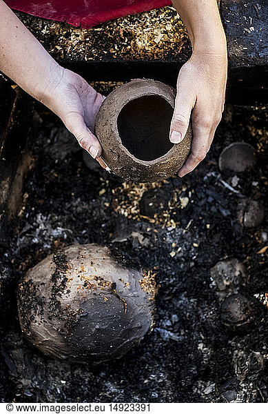 Hochwinkel-Nahaufnahme eines Keramikkünstlers  der neben einer Rauchfeuergrube im Freien sitzt und an einer Vase arbeitet.