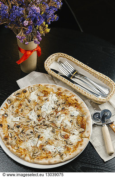 Hochwinkelansicht einer Pizza im Teller mit Besteck und Serviette bei einer Blumenvase auf dem Tisch