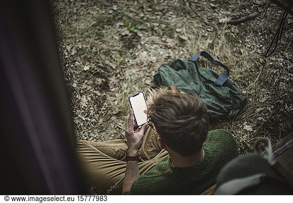 Hochwinkelansicht eines Mannes  der im Schneidersitz sitzt  während er ein Smartphone gegen ein Wohnmobil beim Camping benutzt