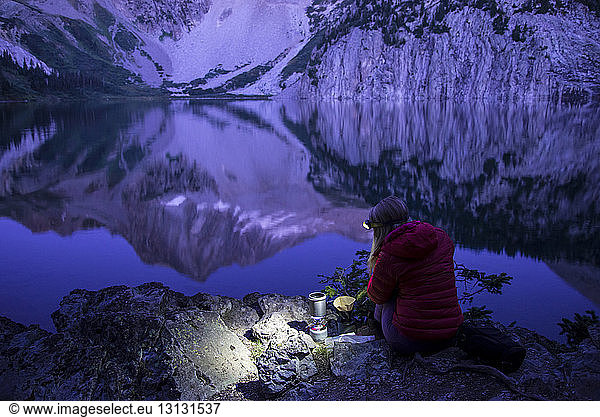 Hochwinkelaufnahme einer Wanderin mit Scheinwerfer beim Kaffeekochen am See vor den Bergen