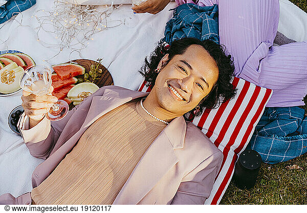 Hochwinkliges Porträt eines lächelnden schwulen Mannes  der ein Weinglas hält  während er sich im Hinterhof hinlegt