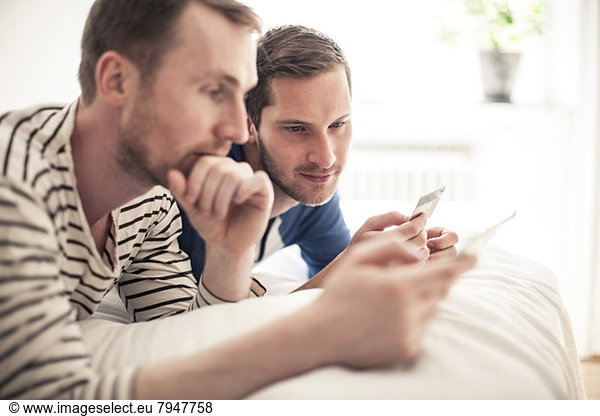 Homosexuelles Paar  das zu Hause im Bett liegt und sich die Kreditkarte ansieht.
