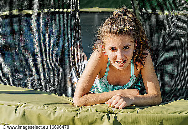 Horizontal portrait of a curly brunette teenager very happy smiling while lying on a trampoline looking at camera. The girl feels happy and tired after jumping on the trampoline.