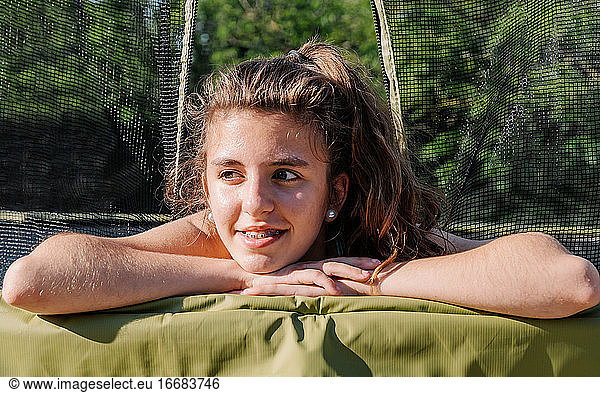 Horizontal portrait of a curly brunette teenager very happy smiling while lying on a trampoline with the net at the background. The girl feels happy and tired after jumping on the trampoline.