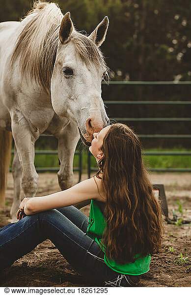 Horse leaning down kissing young teen sitting on ground