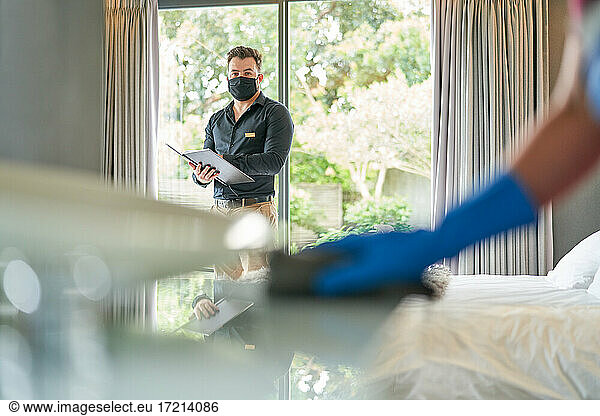 Hotel manager in face mask inspecting room and watching maid clean