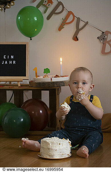 Hungriger kleiner Junge  der den Kuchen seines ersten Geburtstags isst  während er gegen die Geburtstagsdekoration zu Hause sitzt