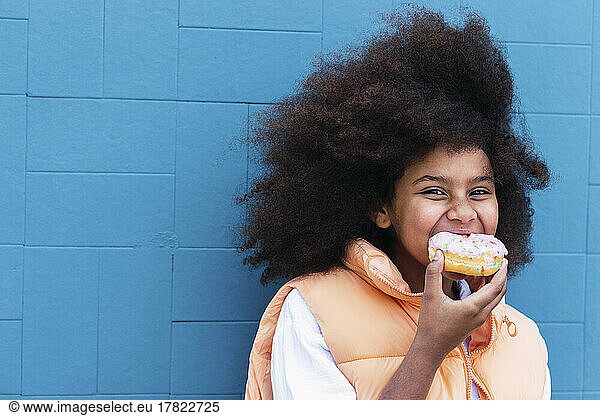 Hungry girl eating doughnut standing in front of blue wall